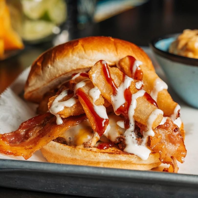 A loaded burger with bacon, cheese, fries, ketchup, and ranch dressing on a bun.