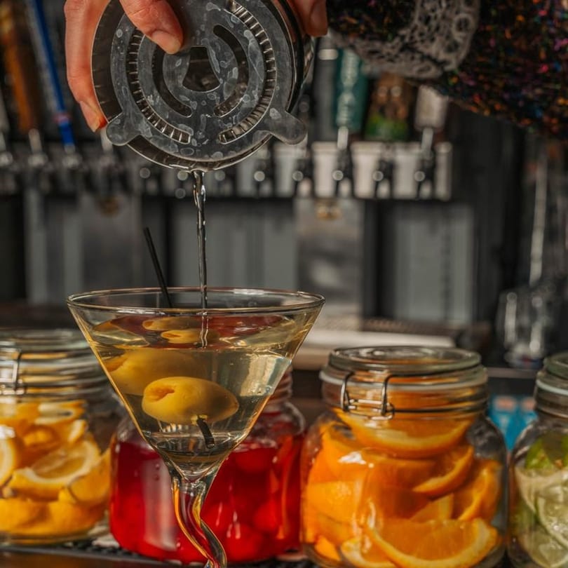 Bartender pouring a martini with olives into a glass, with jars of fruit slices in the foreground.