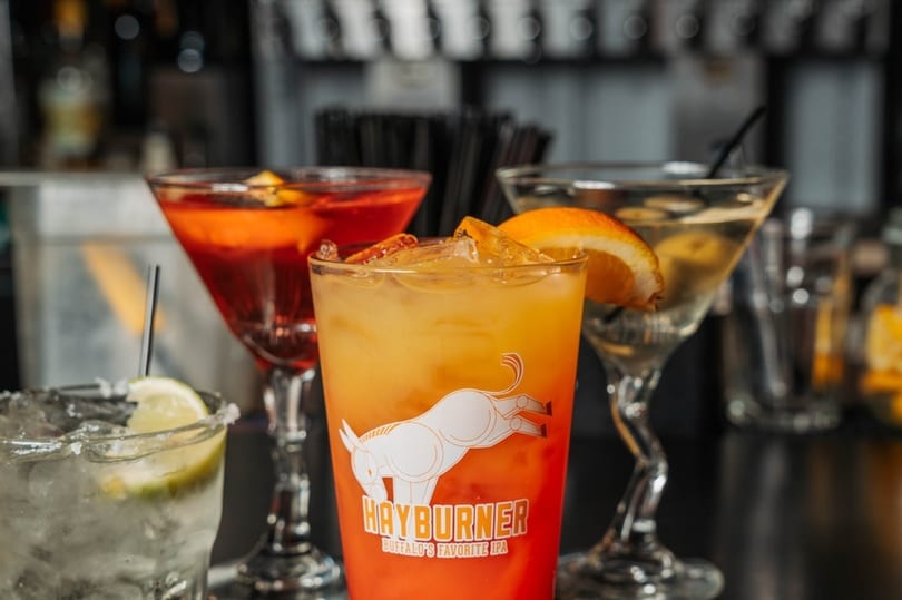 Four colorful cocktails, including a Hayburner IPA glass, on a dark bar surface.