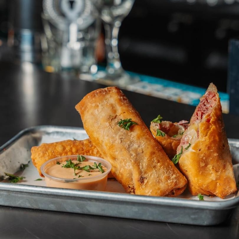 Crispy golden-brown egg rolls, one cut open to reveal corned beef and cheese, with dipping sauce.