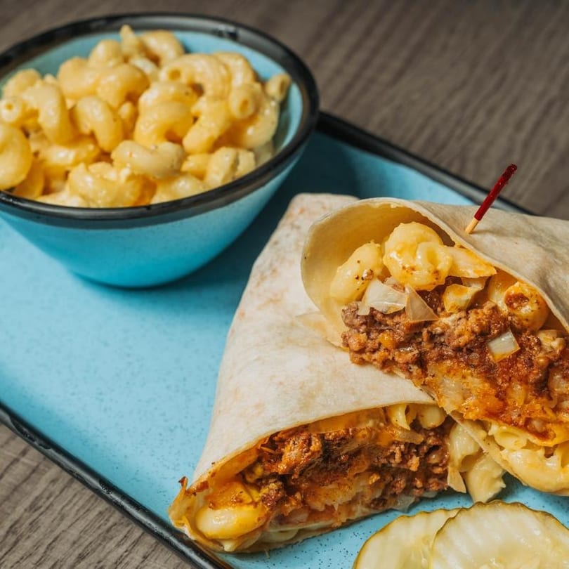 A chili mac and cheese burrito, cut in half, with a side of mac and cheese and pickles.